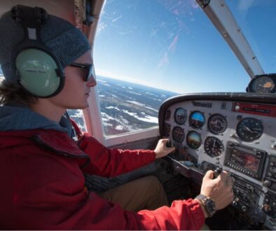 Pilot wearing headset and sunglasses operating controls in cockpit with scenic snowy landscape outside, showing hands-on management of slip vs skid using turn coordinator ball.