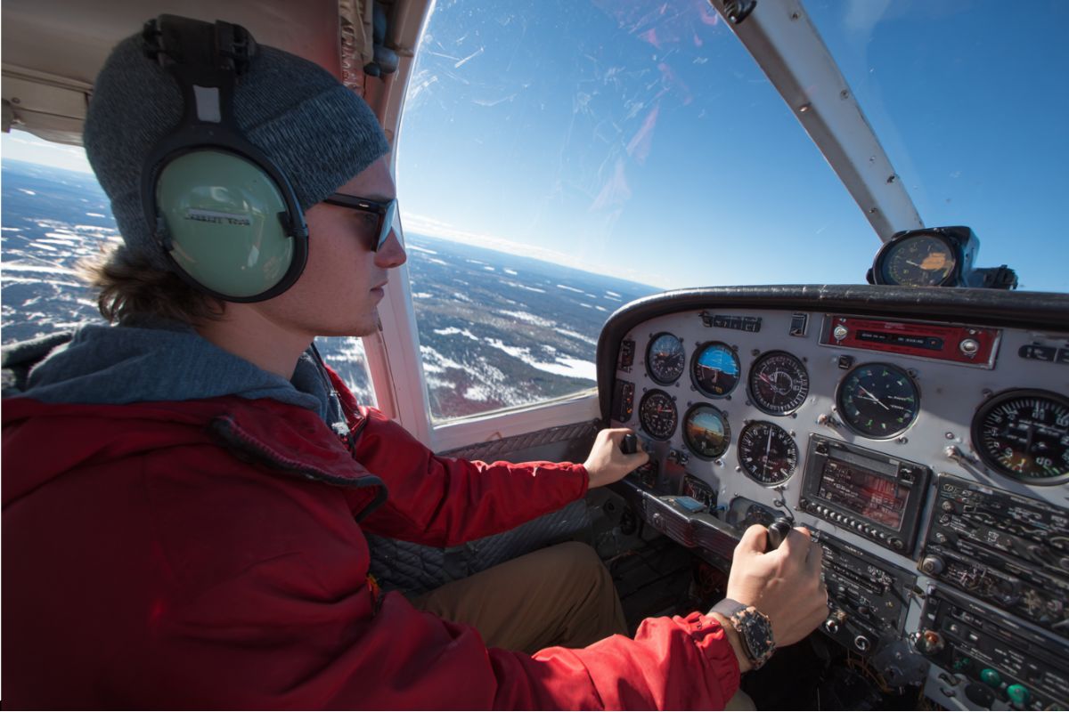 Pilot wearing headset and sunglasses operating controls in cockpit with scenic snowy landscape outside, showing hands-on management of slip vs skid using turn coordinator ball.